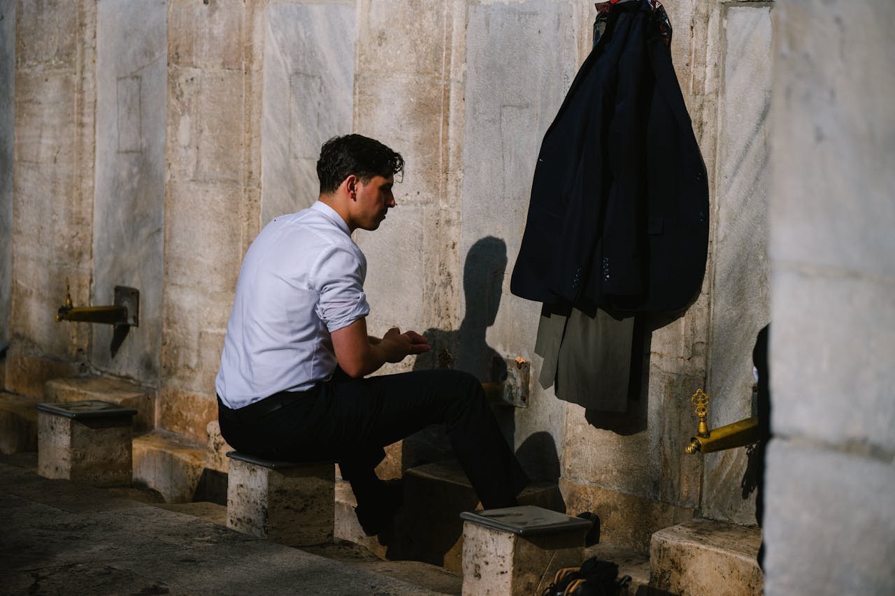 The Art of Drawing Readers In: Your attractive post title goes here A man in a white shirt performing ablution at an outdoor water faucet, with clothes hung nearby.
